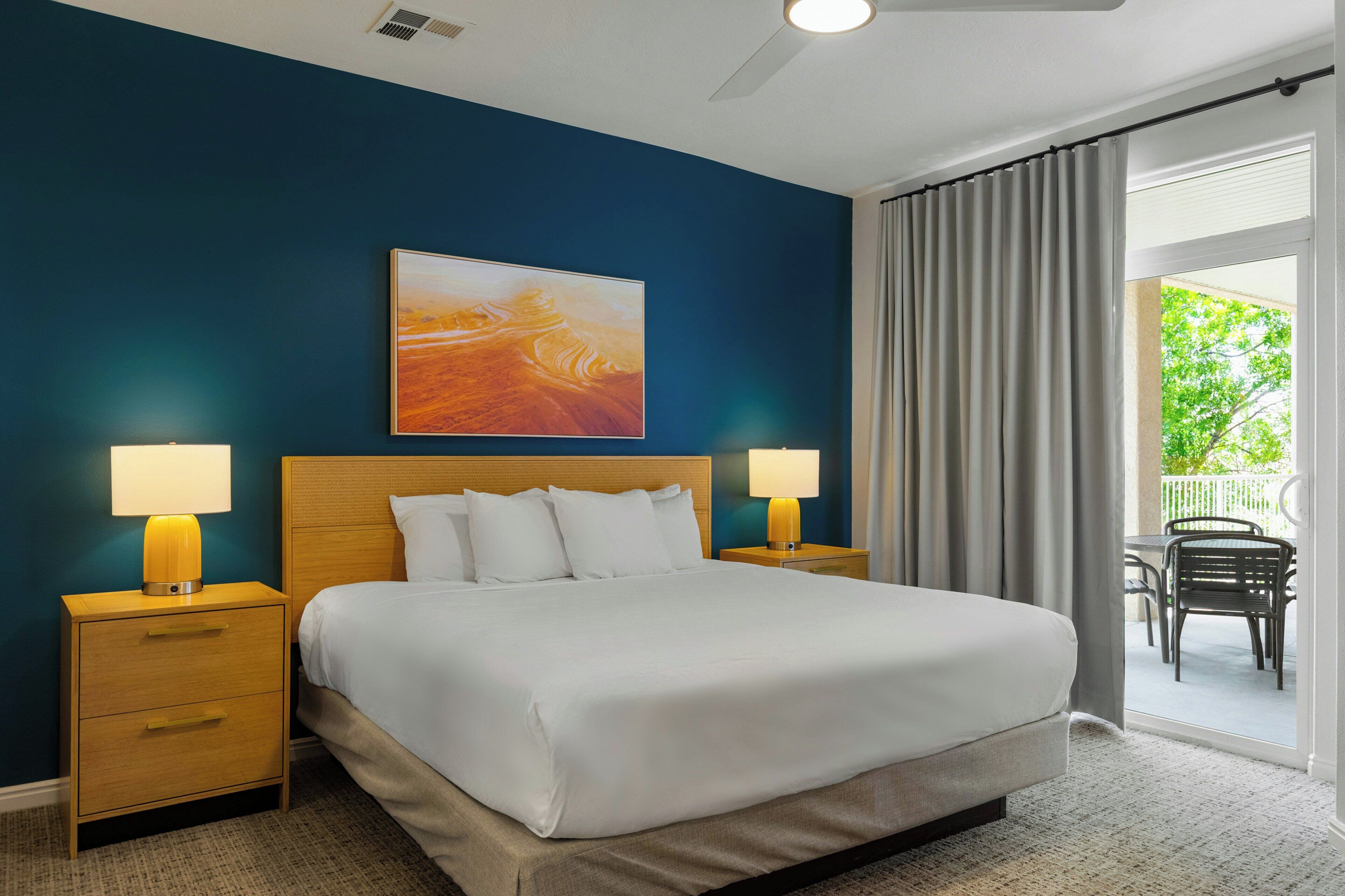A hotel room with a large bed, two bedside tables with lamps, a painting on the wall, and a view of the outdoors through a sliding door.