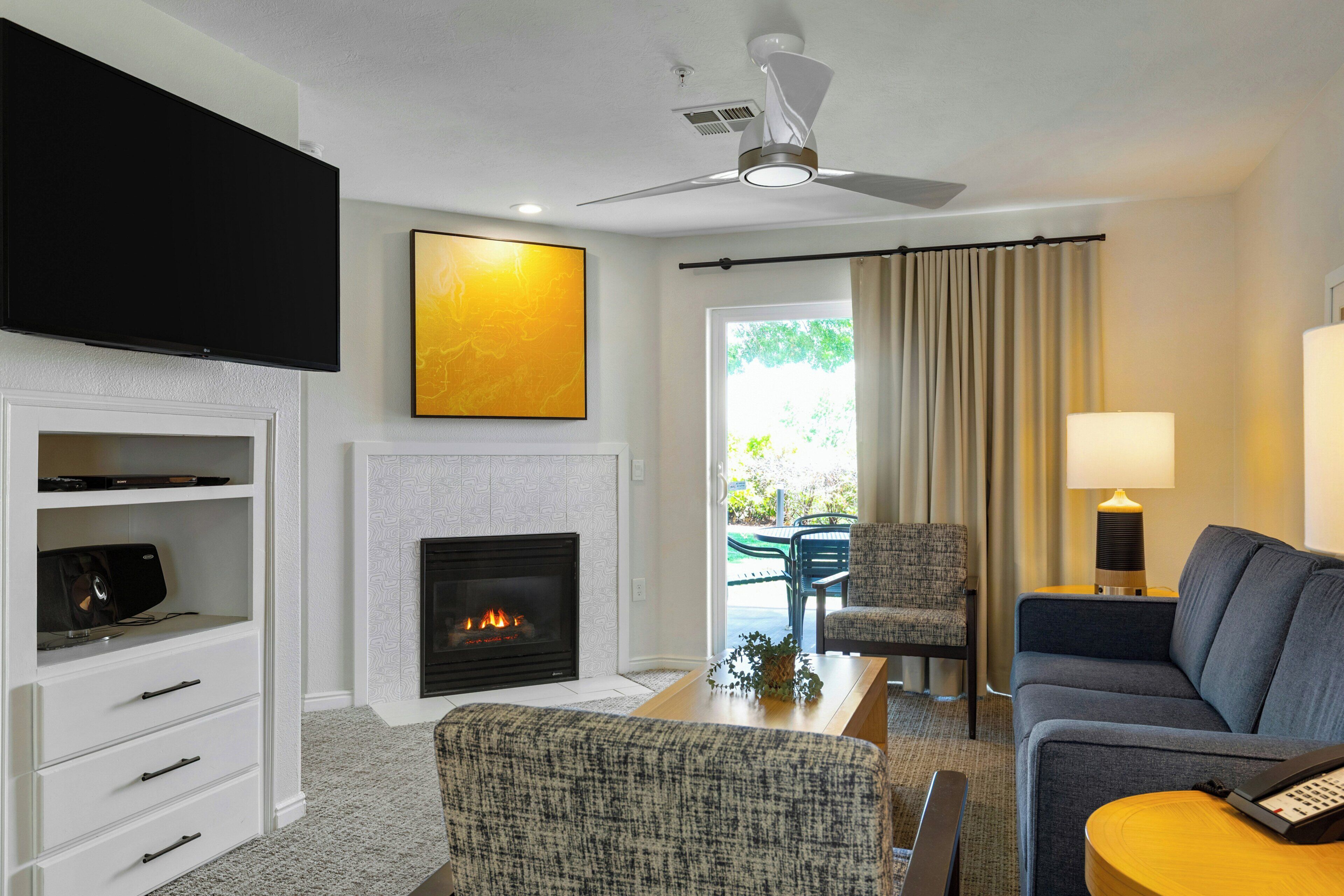 A modern living room with a flat-screen TV, a fireplace, a ceiling fan, and a view of the outdoors.