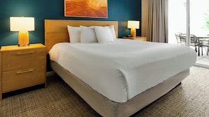 A hotel room with a large bed, two bedside tables with lamps, a wooden headboard, a painting above the bed, and a view of the outdoors through a window. - WorldMark St. George (St. George)