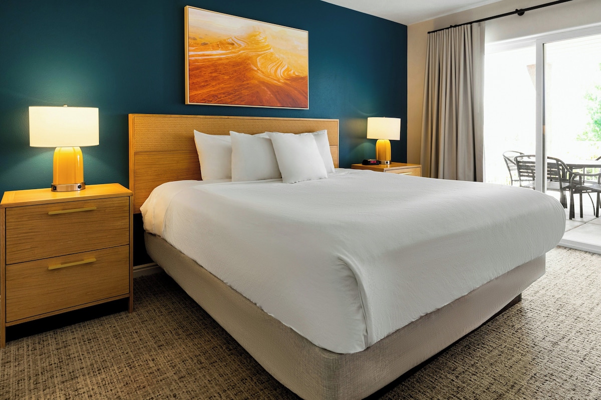 A hotel room with a large bed, two bedside tables with lamps, a wooden headboard, a painting above the bed, and a view of the outdoors through a window.