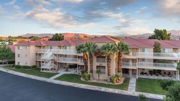 A multi-story residential building with balconies, palm trees, and a view of mountains in the background.