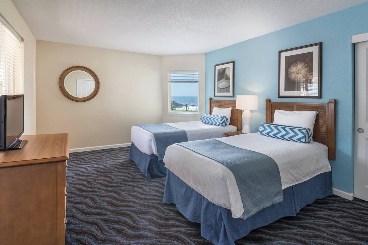 A hotel room with two beds, a desk, a mirror, and a window with a view.