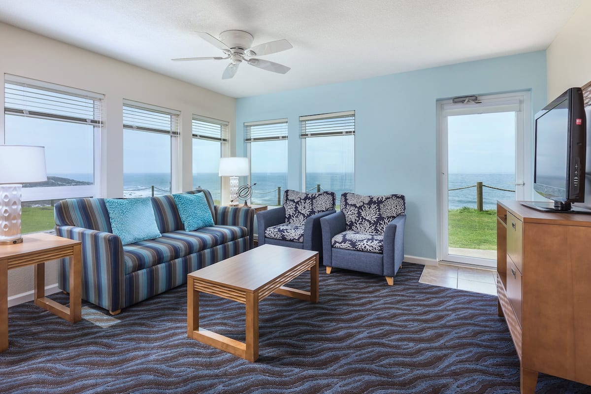 A living room with a sofa, two armchairs, a coffee table, a TV, and a view of the ocean through large windows.
