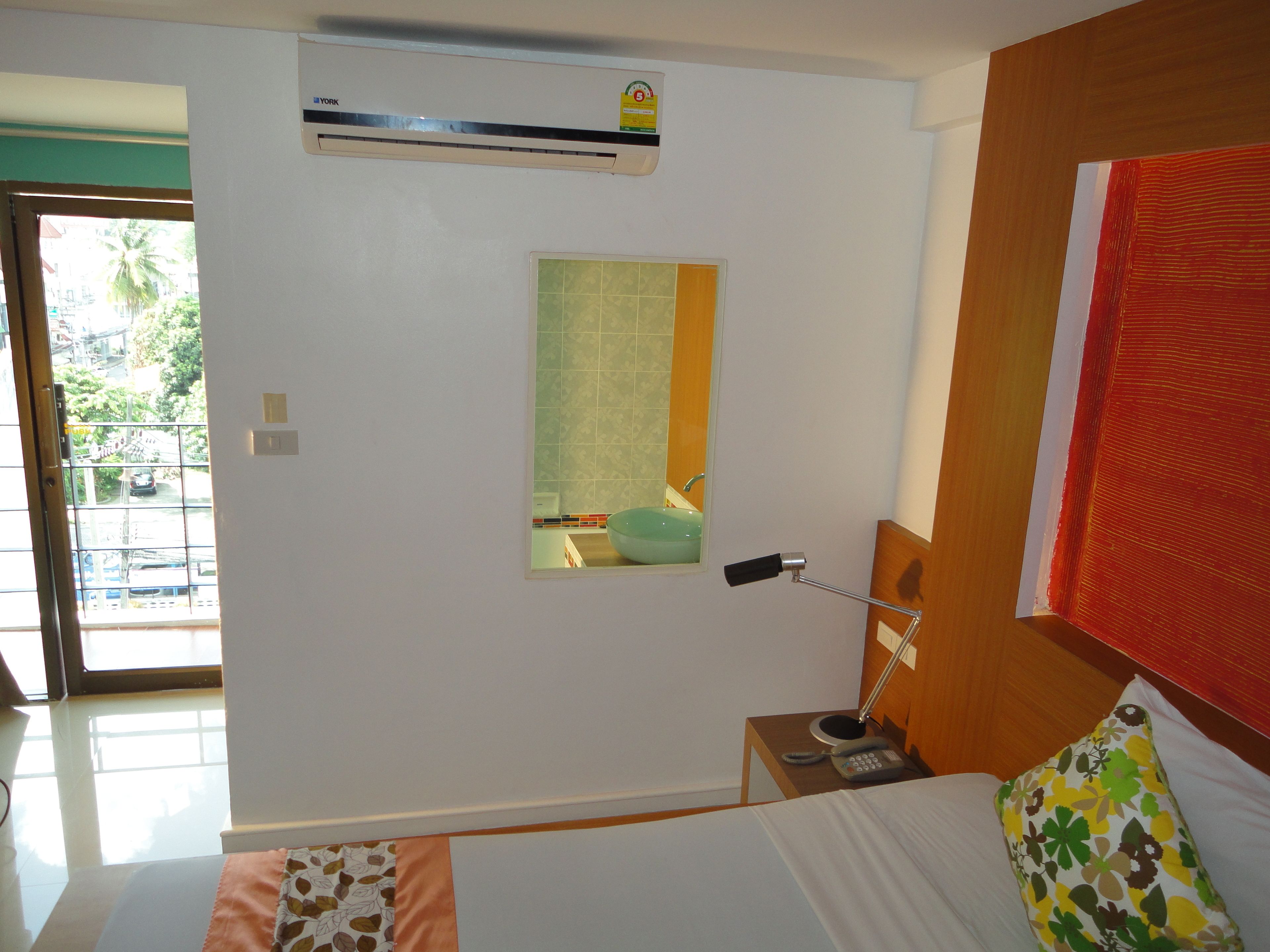 A hotel room with a bed, a bedside table with a lamp, a small window, and an air conditioning unit on the wall.
