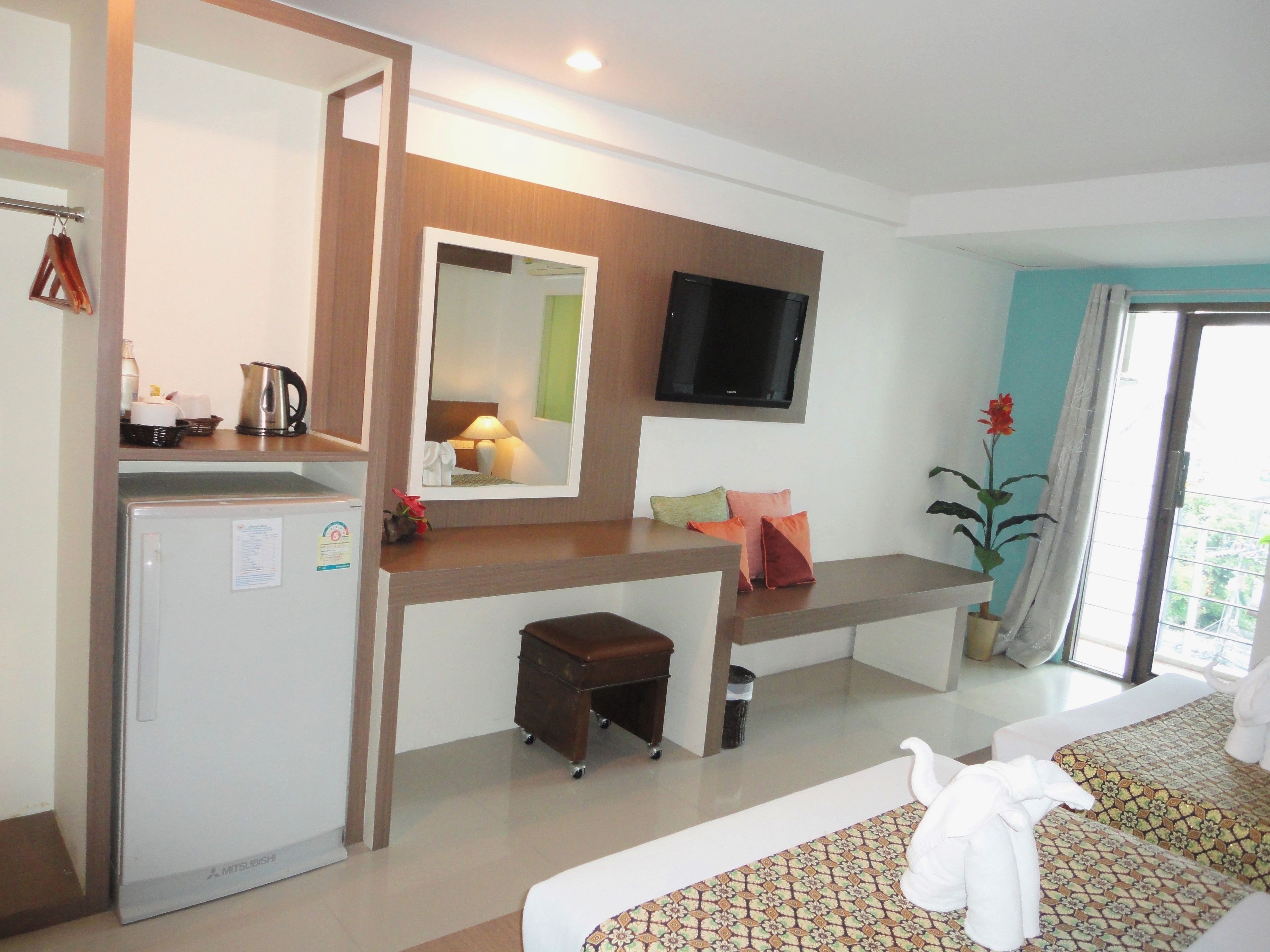 A hotel room with a bed, a desk with a mirror, a television, a refrigerator, and a small stool.