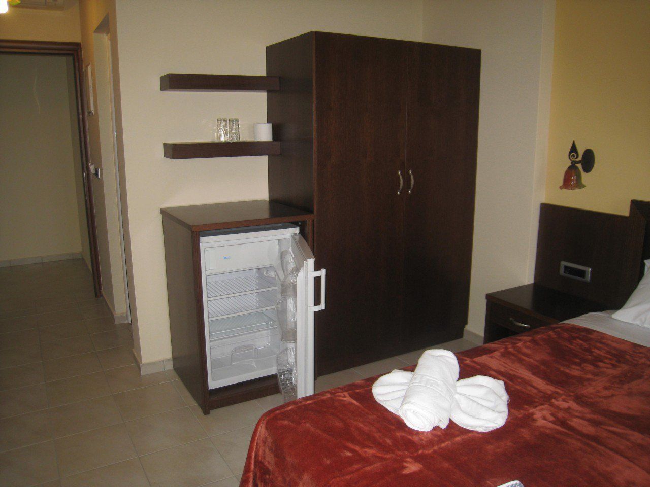 Une chambre d’hôtel avec un lit, une armoire, un minibar et une petite table avec une bouteille.