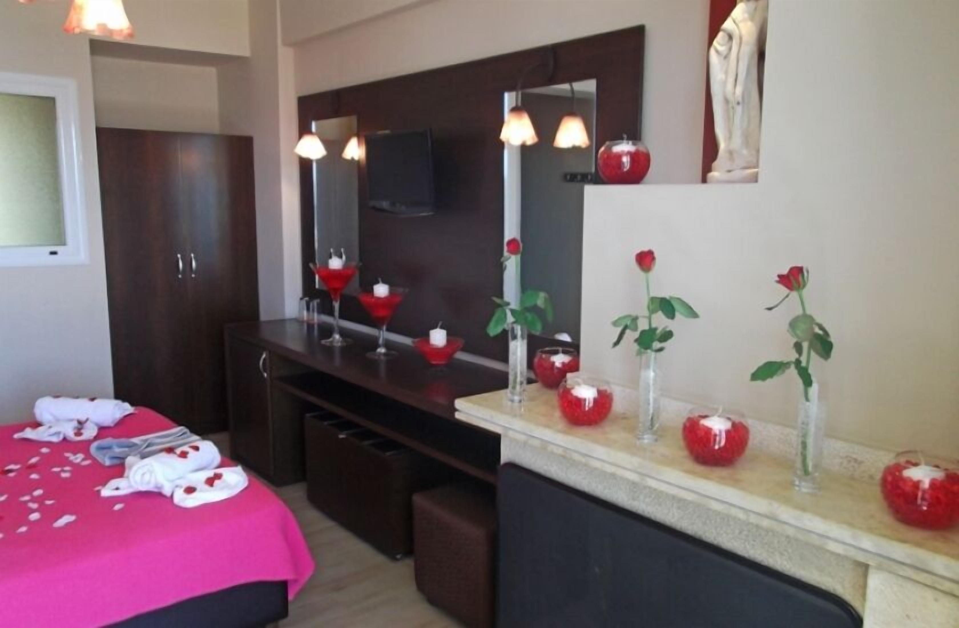 Habitación de hotel con cama, tocador con espejo y cómoda. Se observan objetos decorativos sobre la cómoda, como rosas rojas y velas.