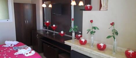 Habitación de hotel con cama, tocador con espejo y cómoda. Se observan objetos decorativos sobre la cómoda, como rosas rojas y velas.
