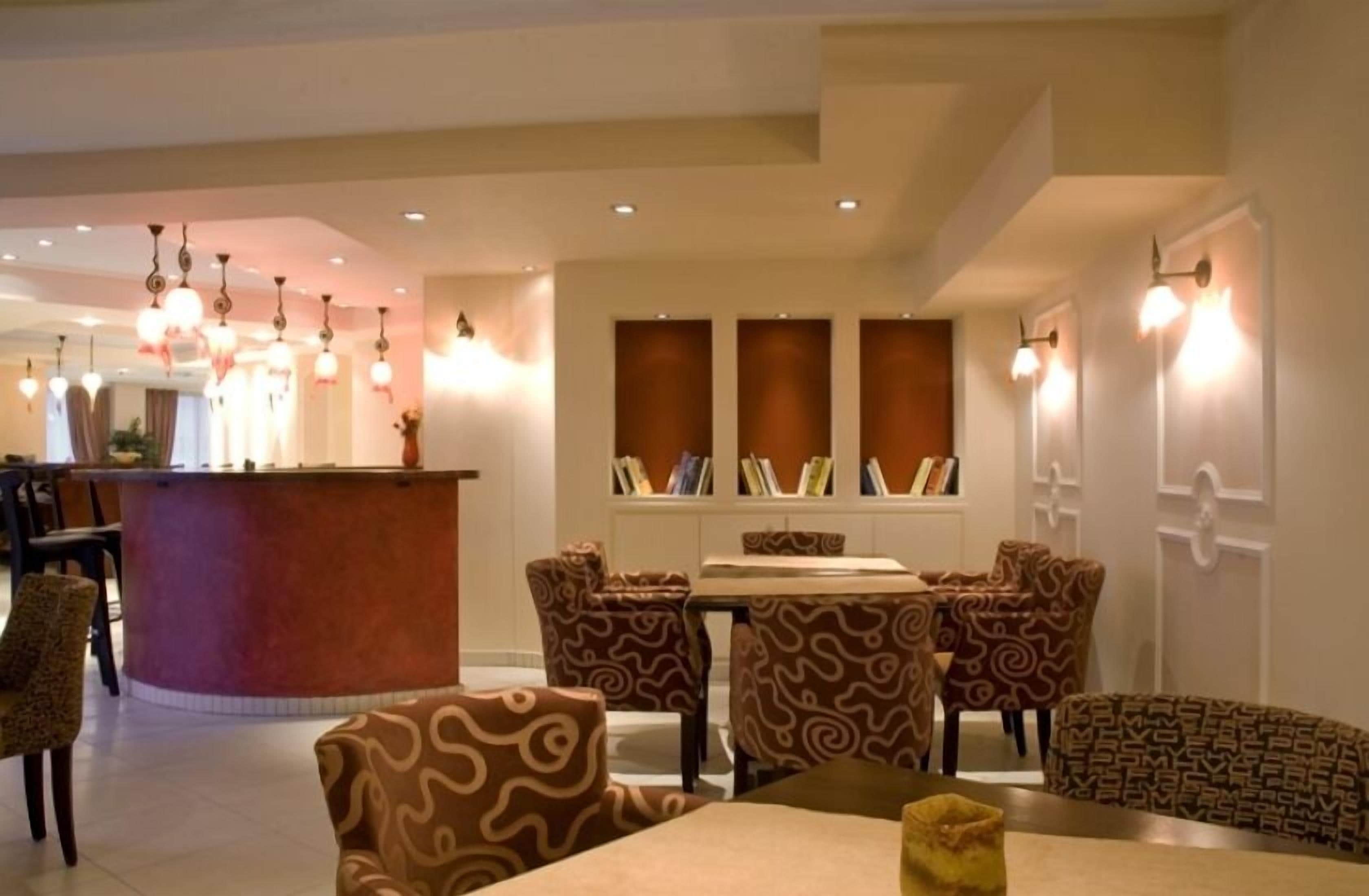 A hotel lobby with a reception desk, patterned chairs, a dining table, and bookshelves.