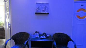 A room with two wicker chairs, a small table with a plate and a bottle, and a wall-mounted shelf with a framed picture.