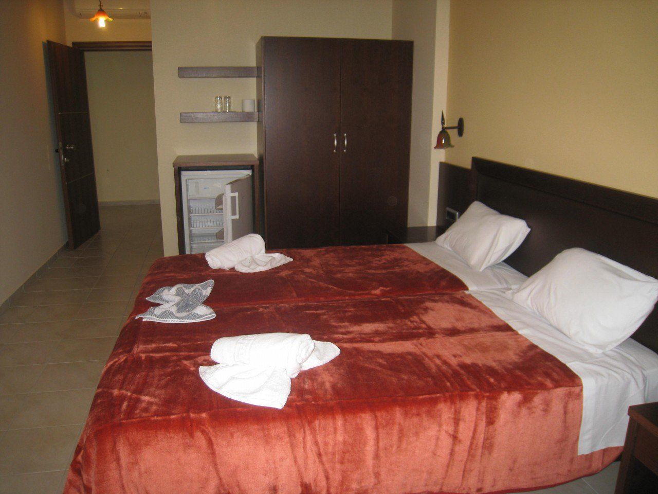 A hotel room with a large bed, a wardrobe, a small desk, and a shelf with toiletries.