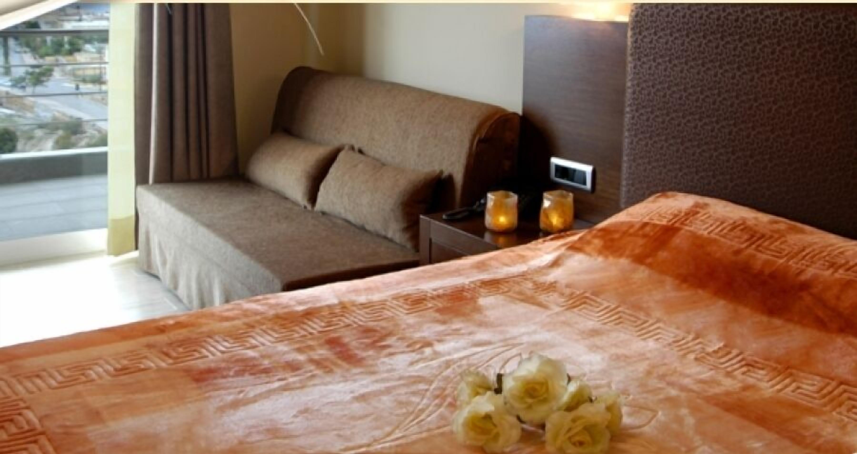 A hotel room with a bed, a sofa, a nightstand with a lamp, and a view of the outdoors.