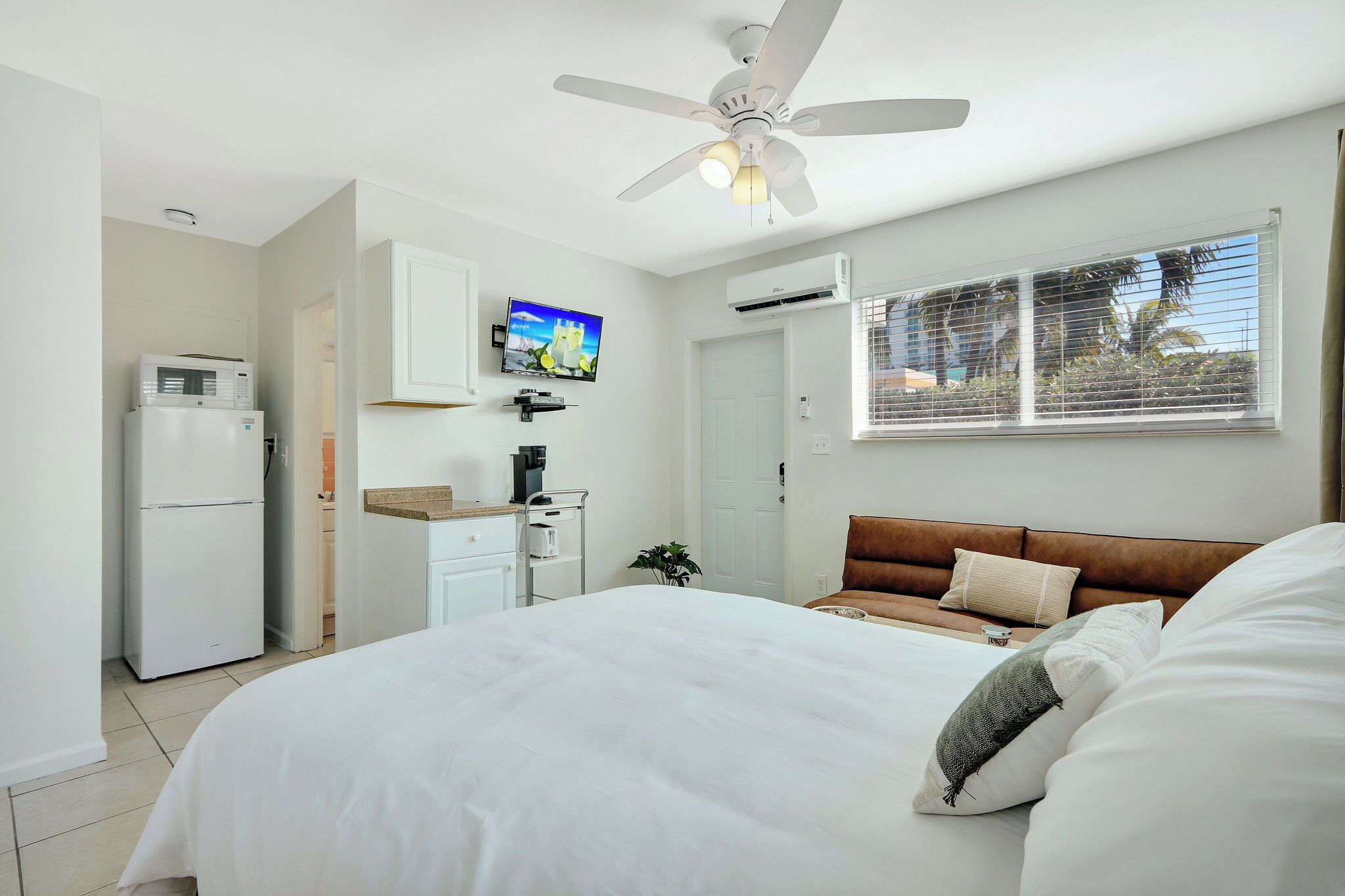 A bedroom with a bed, a ceiling fan, a TV, a refrigerator, and a window with blinds.