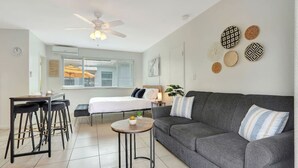 A modern living room with a grey sofa, a round coffee table, a bed with white bedding, a ceiling fan, and decorative wall art. - Dream Inn Hotel (Hollywood)