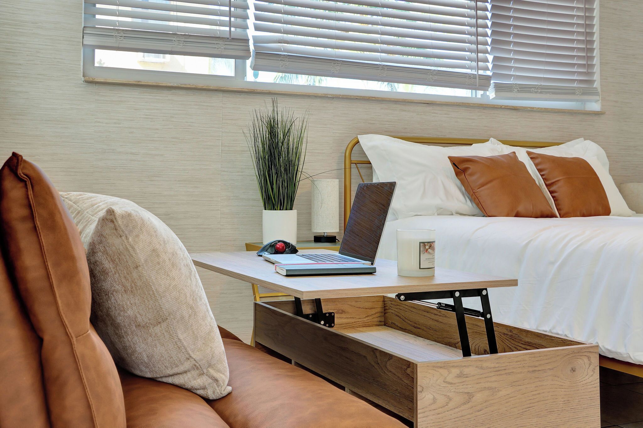 A modern bedroom with a bed, a desk with a laptop, a potted plant, and a window with blinds.