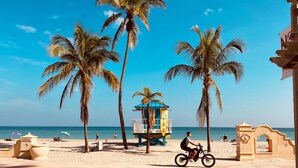 A beach scene with palm trees, a lifeguard tower, and a person riding a bicycle. - Dream Inn Hotel (Hollywood)