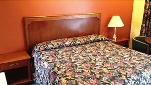 A hotel room with a wooden headboard, a floral bedspread, a nightstand with a lamp, and a small drawer. - Parkway Motel (London)