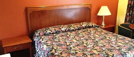 A hotel room with a wooden headboard, a floral bedspread, a nightstand with a lamp, and a small drawer.