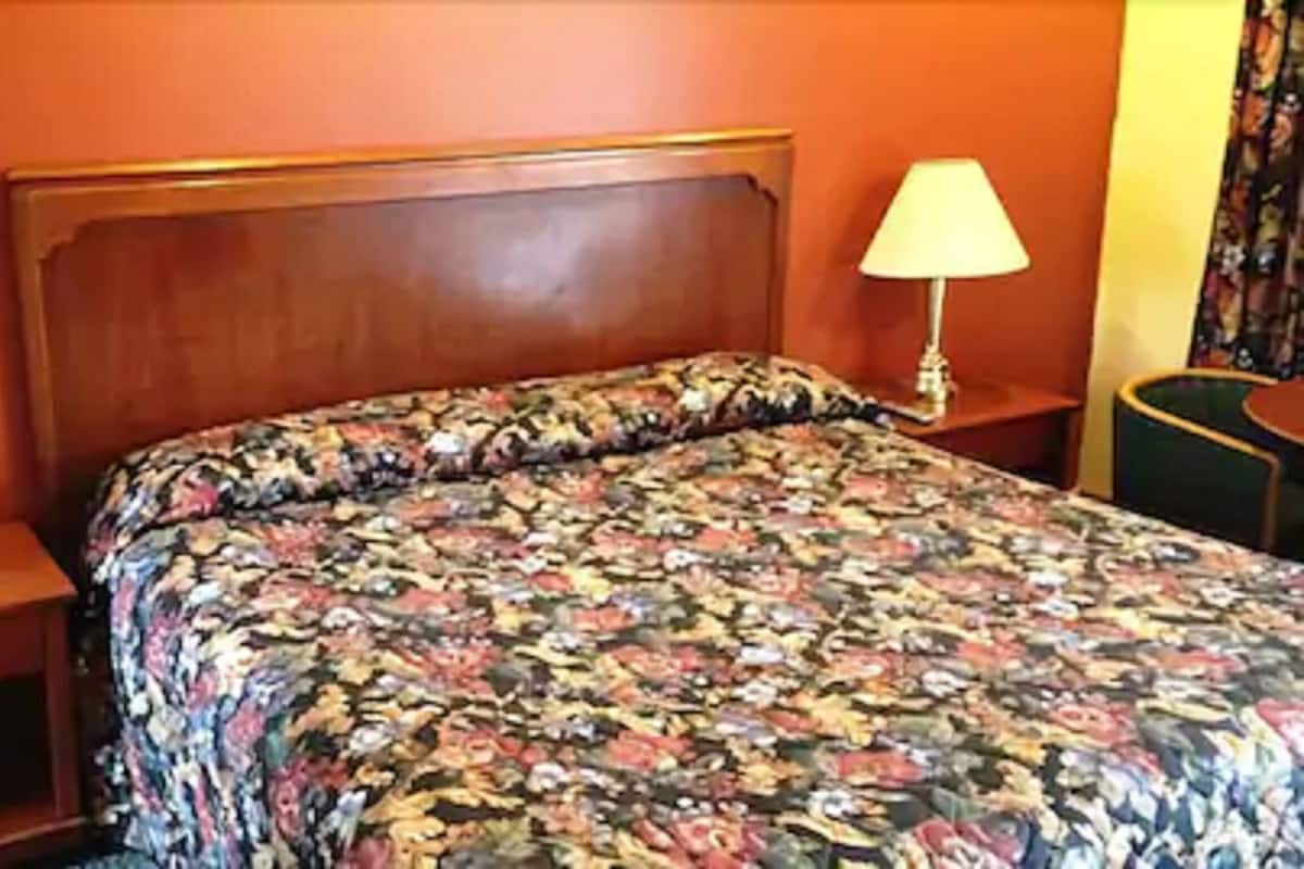 A hotel room with a wooden headboard, a floral bedspread, a nightstand with a lamp, and a small drawer.