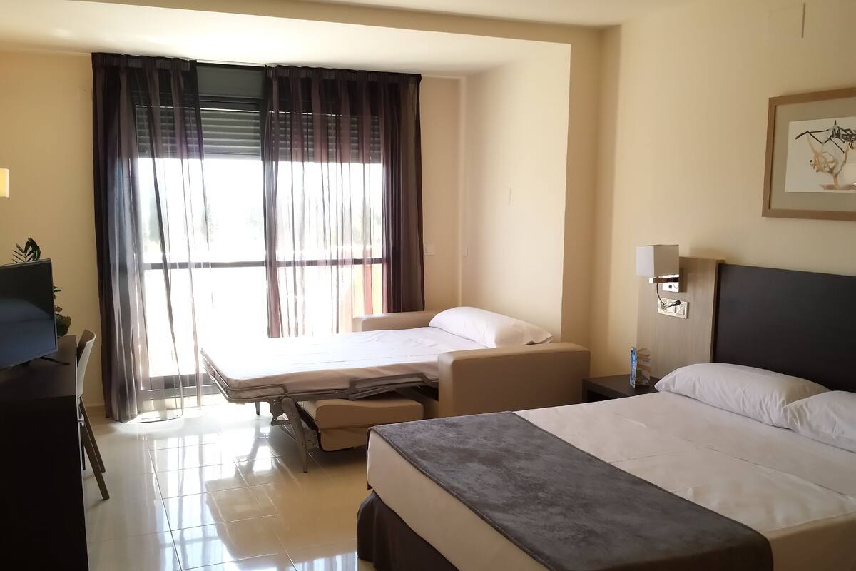 A hotel room with a bed, a desk, a chair, a TV, and a window with curtains.