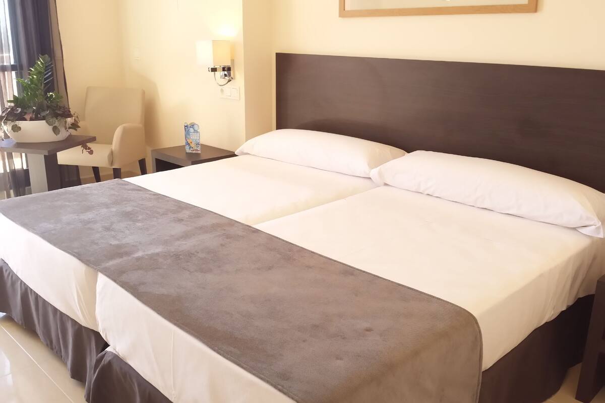 A double bed with a grey headboard and white linens, a bedside table with a lamp, a chair, and a potted plant.