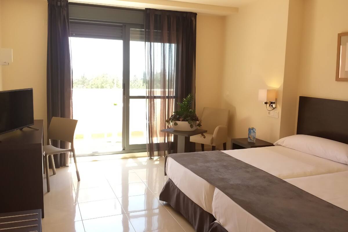 A hotel room with a bed, a desk with a TV, a chair, a small table with a plant, and a view of the outdoors through a sliding door.