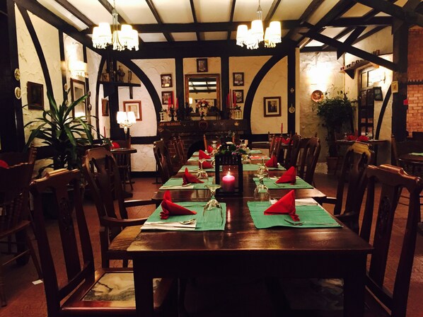 A long dining table set with red napkins, candles, and glassware, surrounded by wooden chairs in a room with exposed beams and framed pictures on the walls.