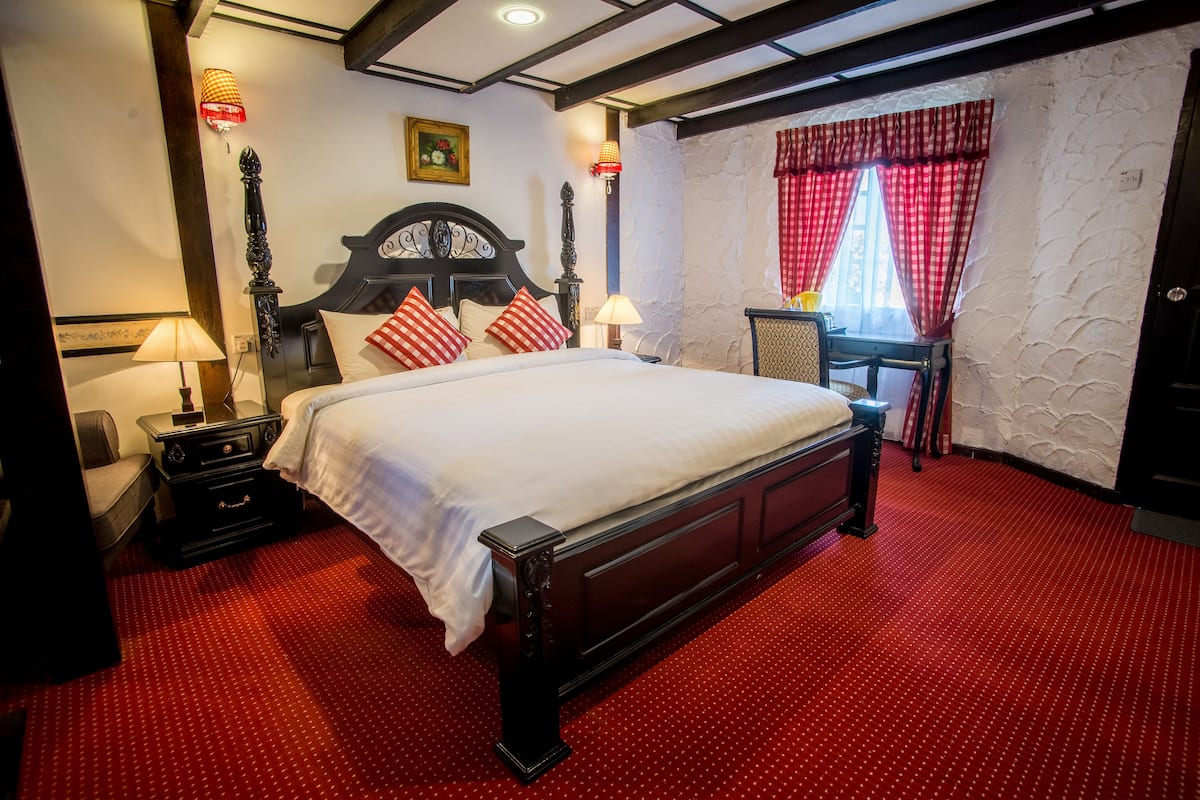A hotel room with a large bed, two bedside tables, a chair, a desk, and a window with red curtains.