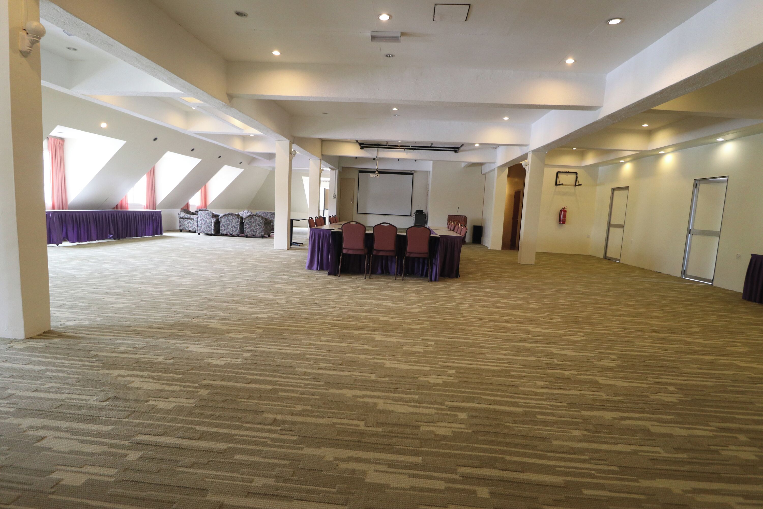 A large, open room with a high ceiling and sloped roof, featuring a long table with chairs, a projector screen, and a fire extinguisher on the wall.