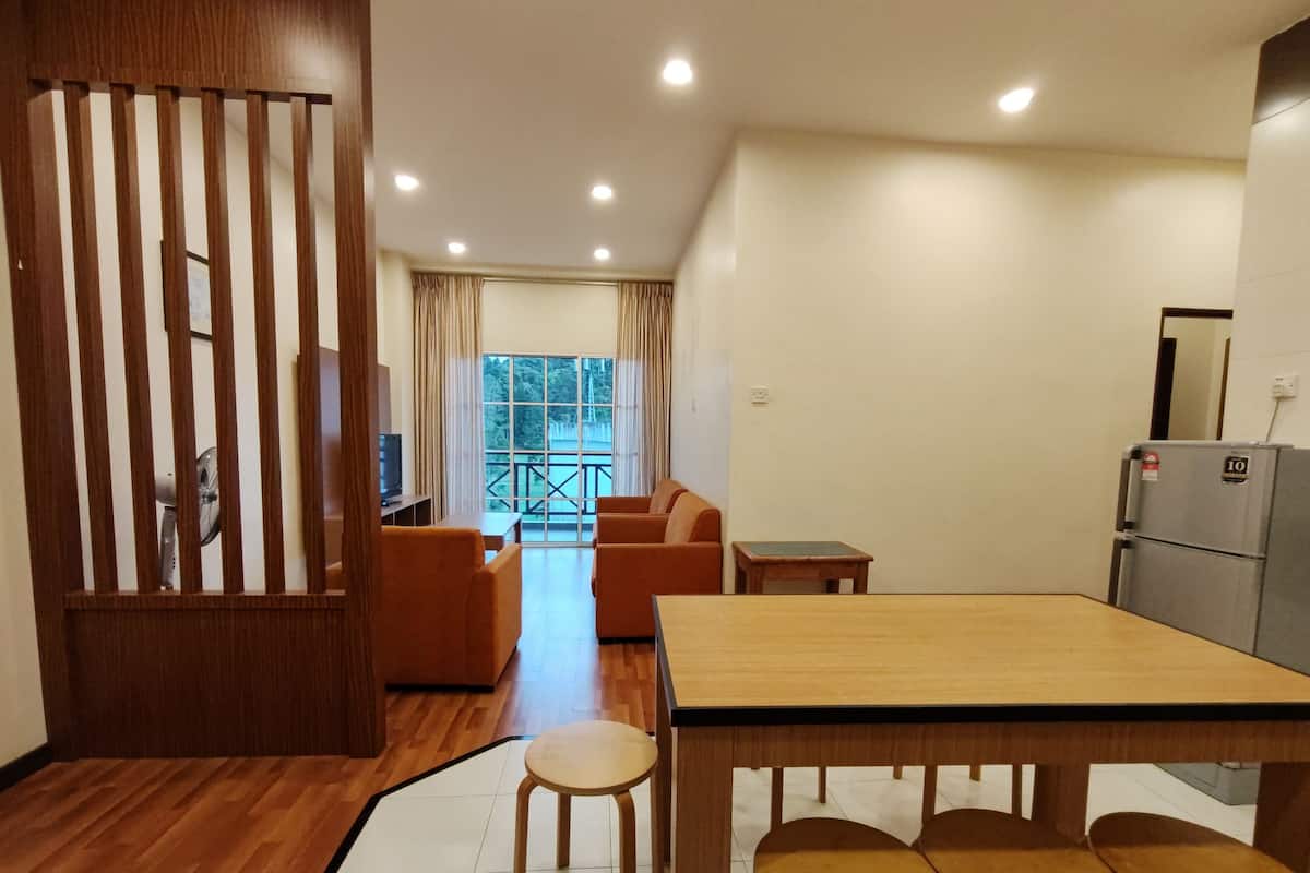 A compact hotel room with a wooden floor, a dining table with four chairs, a sofa, a small table, and a refrigerator.