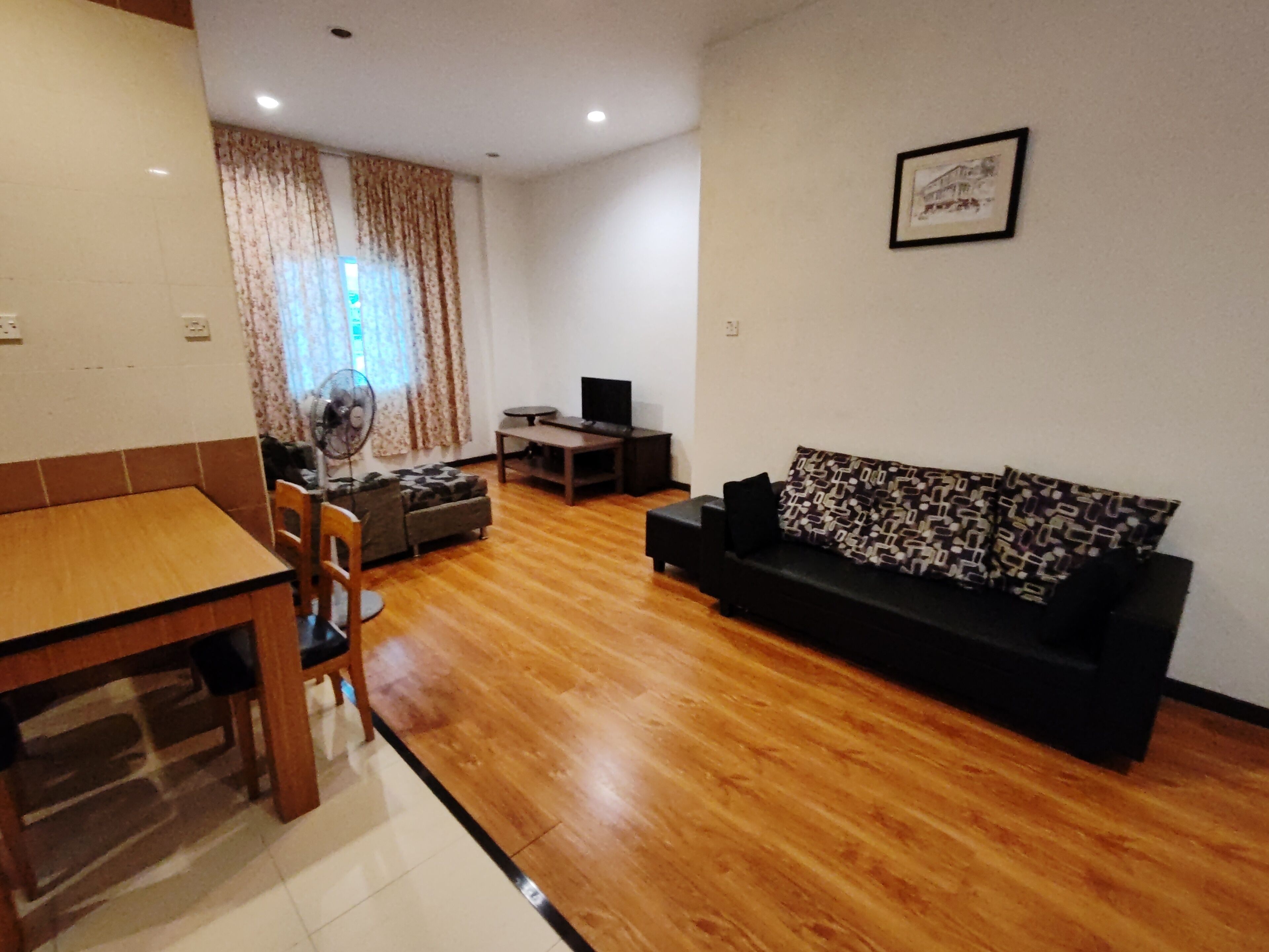 A living room with a wooden floor, a black sofa, a dining table with chairs, a TV, and a fan.