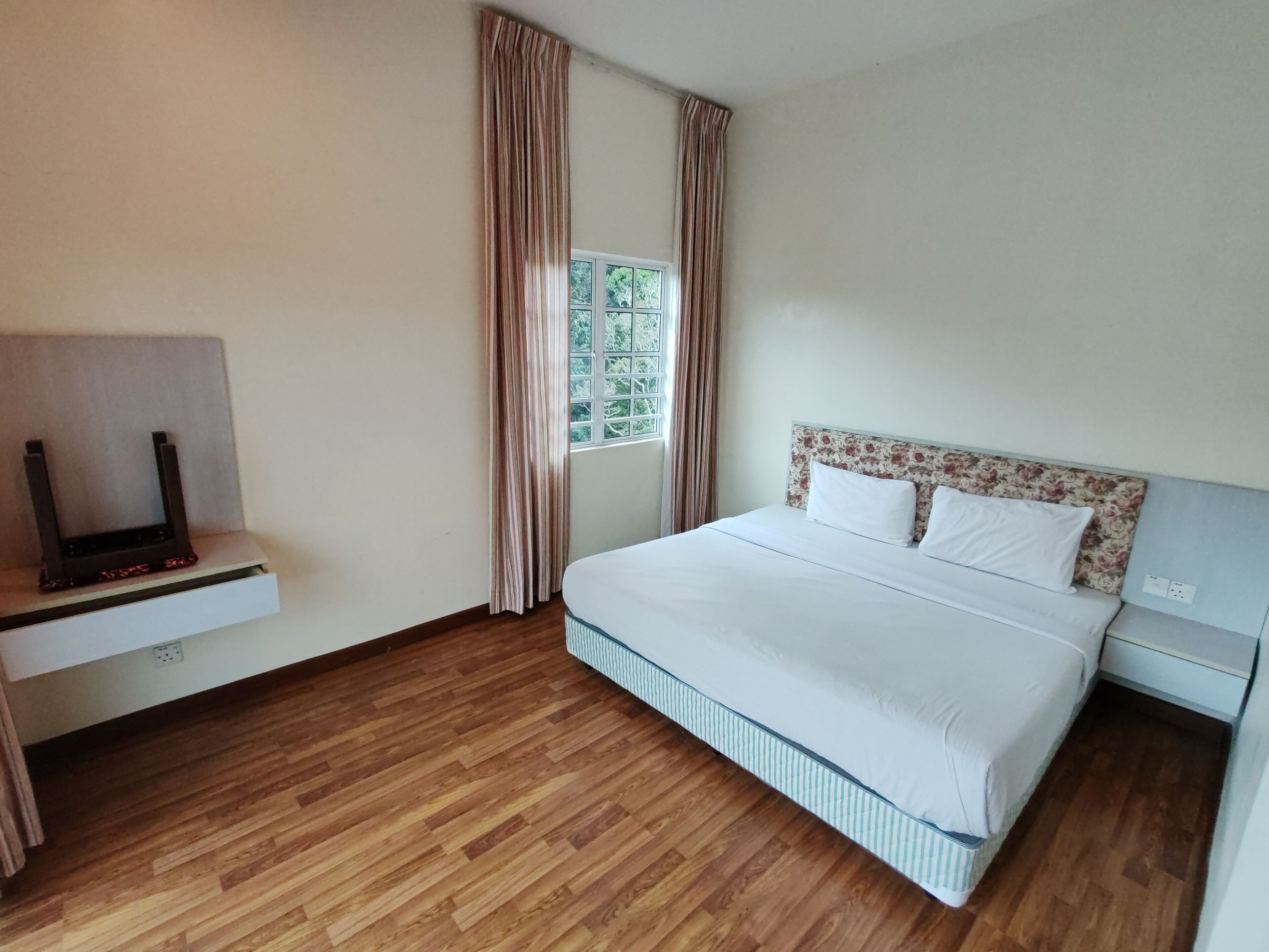 A hotel room with a bed, a TV on a stand, a window with curtains, and a wooden floor.
