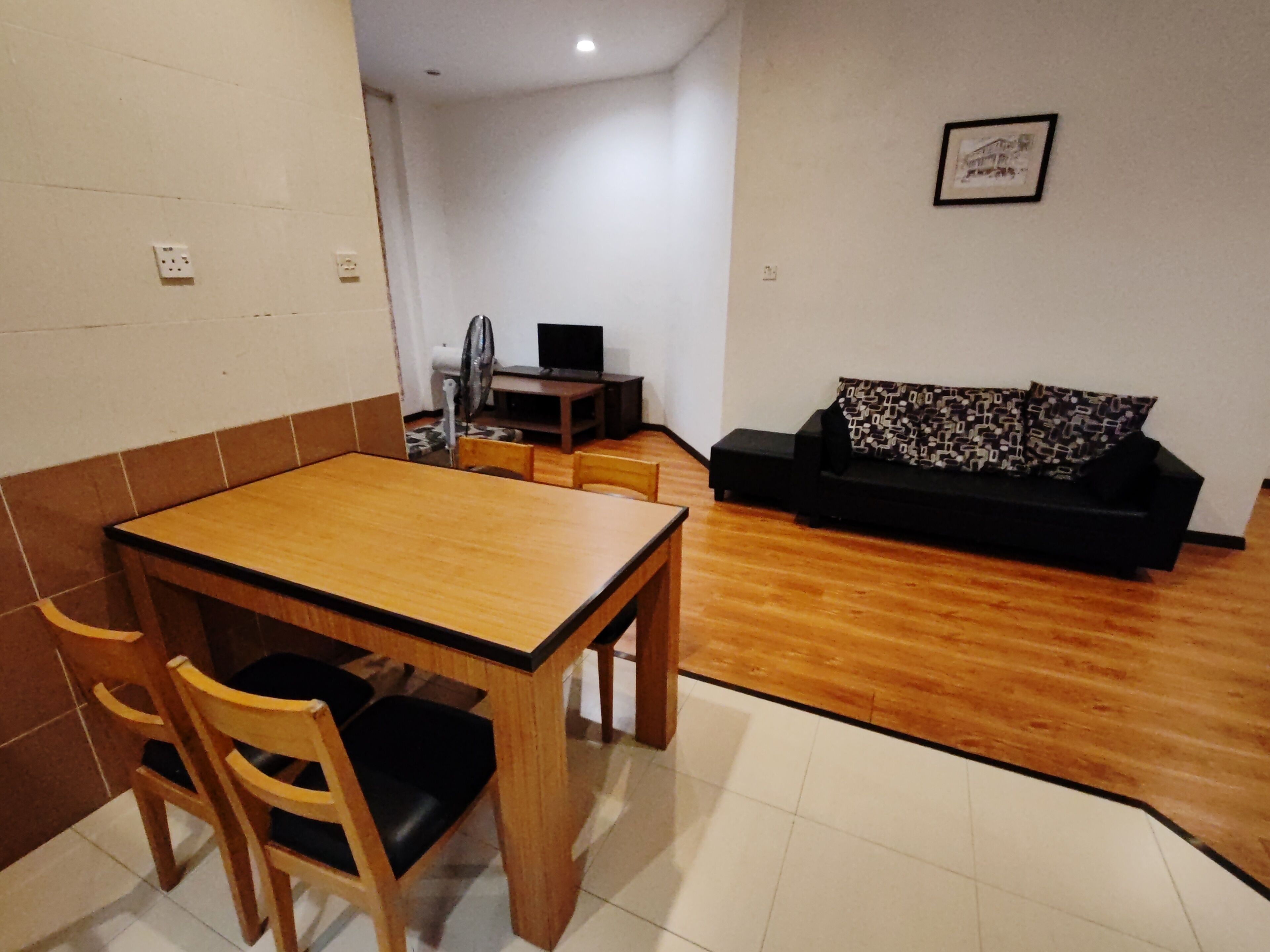 Um quarto pequeno e bem iluminado, com uma mesa de jantar de madeira e cadeiras, um sofá preto com almofadas estampadas e uma televisão pequena sobre um suporte.