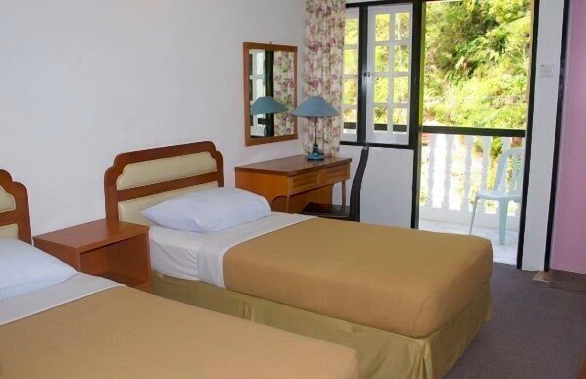 A room with two single beds, a desk with a chair, a mirror, and a window with a view of greenery.