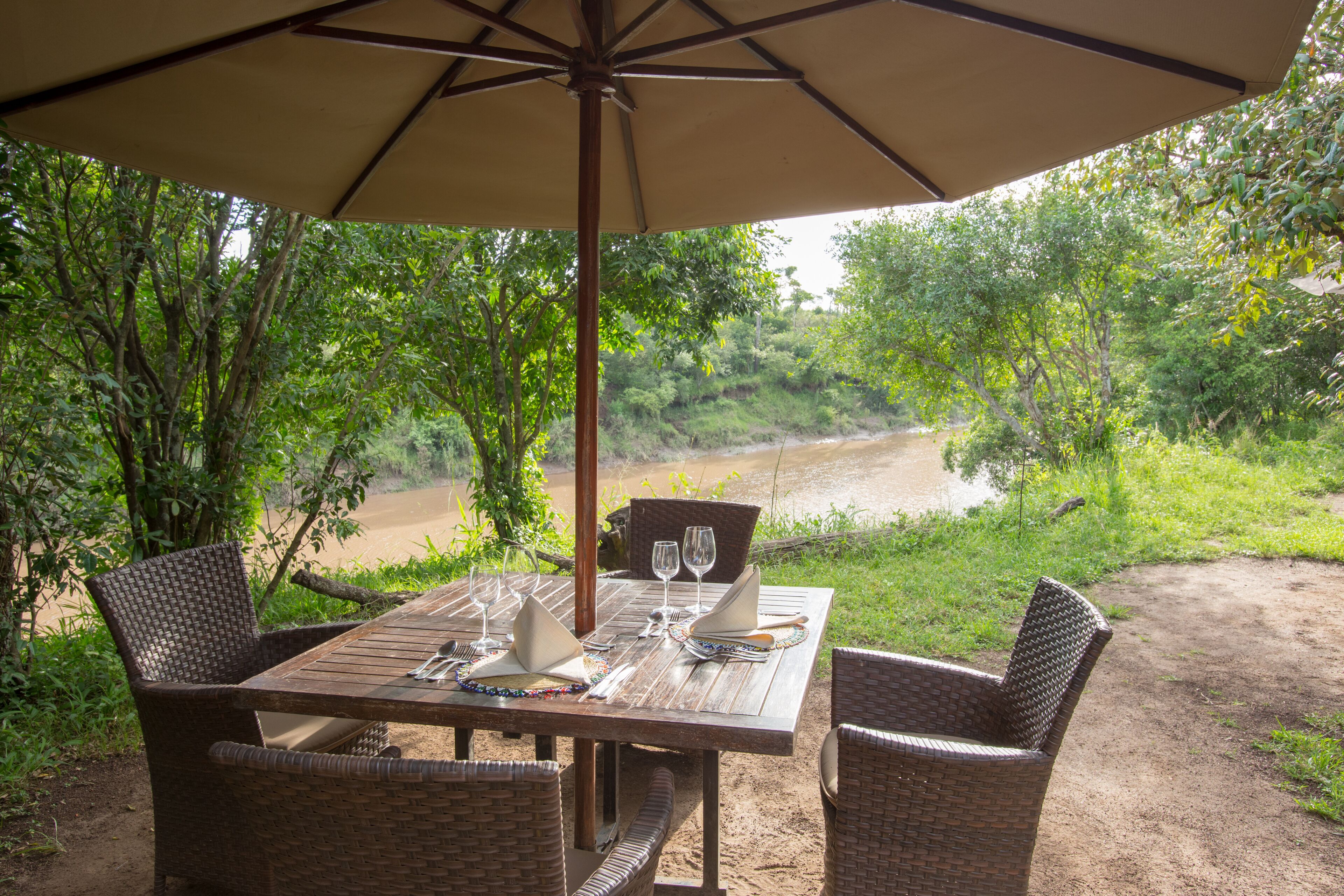 An outdoor dining area with a table set for two, surrounded by wicker chairs, under a large umbrella.