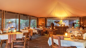 An indoor dining area with tables set for multiple guests, featuring a tent-like roof structure and natural light from large windows.