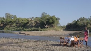 A dining table set up by a river with chairs and a person standing nearby.