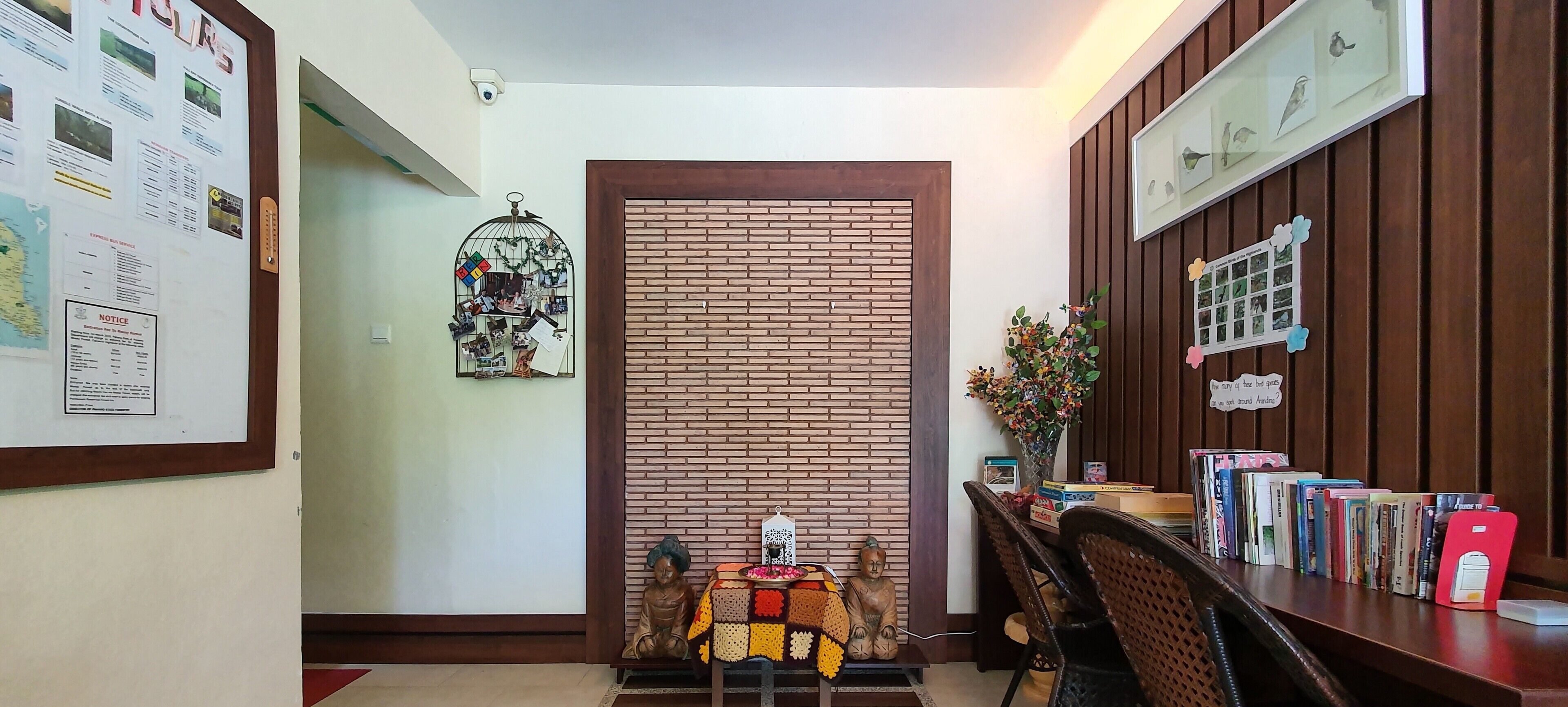 A room with a brick wall, a wooden door, a small table with a statue, a bookshelf with books, and a wall-mounted birdcage with photos.