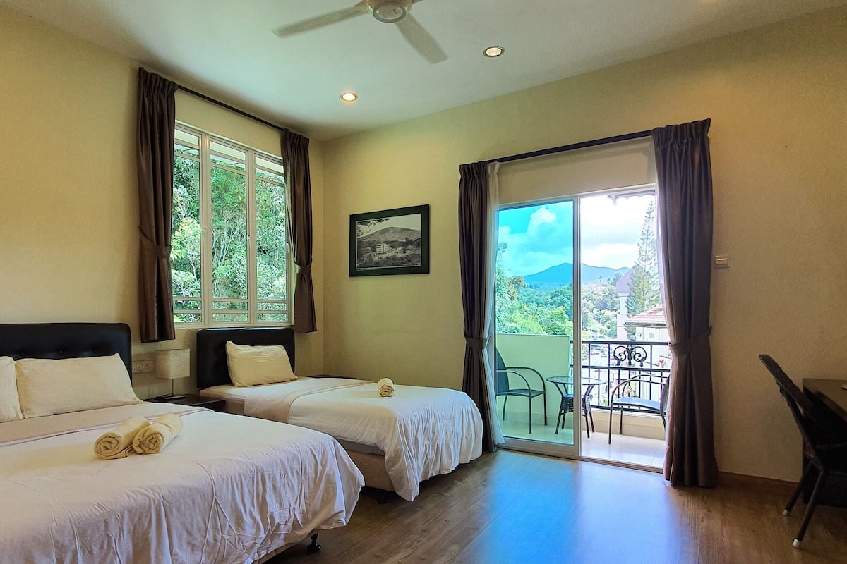 Quarto de hotel com duas camas, uma mesa de trabalho e varanda com vista para vegetação e montanhas.