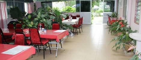 A covered outdoor dining area with red tables and chairs, surrounded by potted plants.