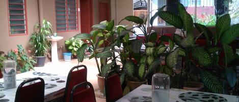A dining area with tables covered in patterned tablecloths, surrounded by potted plants.