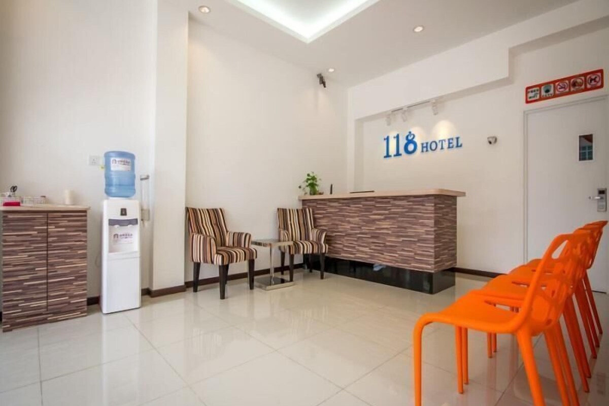 Zona de recepción de un hotel, con mostrador, dos sillones a rayas, dispensador de agua y un cartel con el número del hotel.