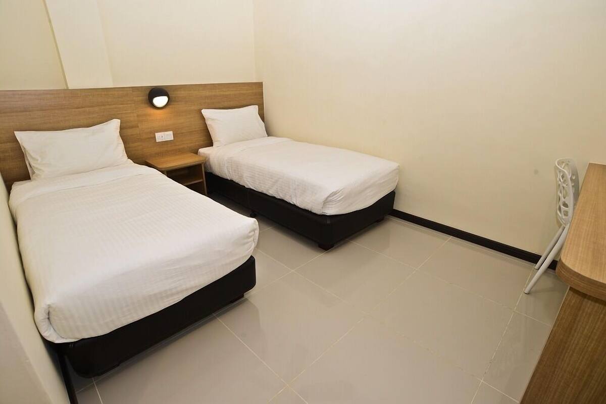 A hotel room with two single beds, a wooden headboard, a bedside table with a lamp, and a small wooden side table.