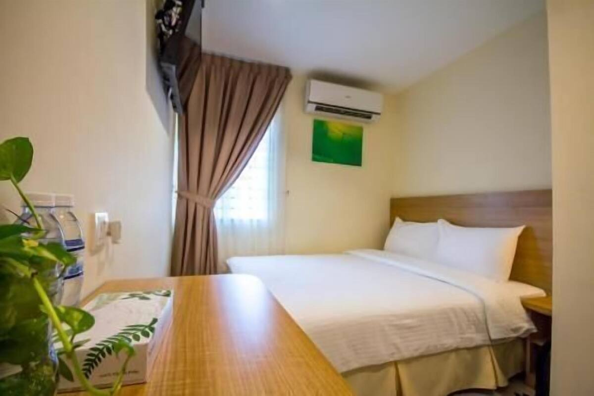A hotel room with a bed, a wooden nightstand, an air conditioning unit, a window with curtains, and a green wall decoration.