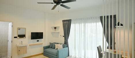 A modern hotel room with a ceiling fan, a flat-screen TV, a sofa, a desk with a chair, and a large window with curtains.