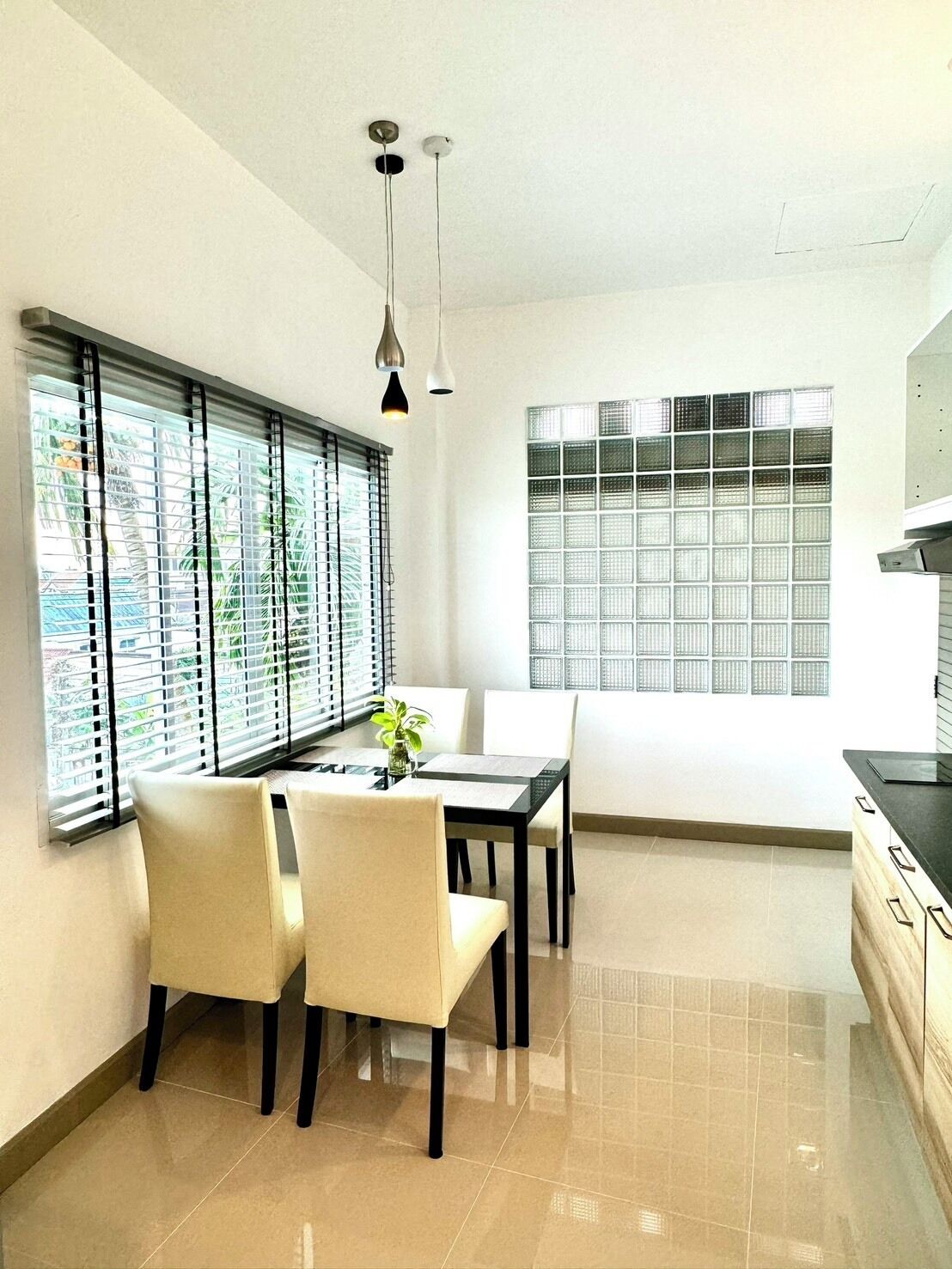 A modern kitchen with a dining area, featuring a large window with blinds, a black countertop, and a hanging light fixture.