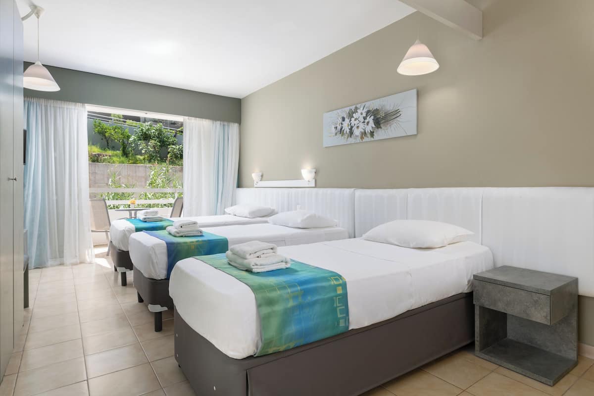 Habitación de hotel con dos camas, una mesita de noche pequeña, un ventanal grande con cortinas y vistas a zonas verdes.