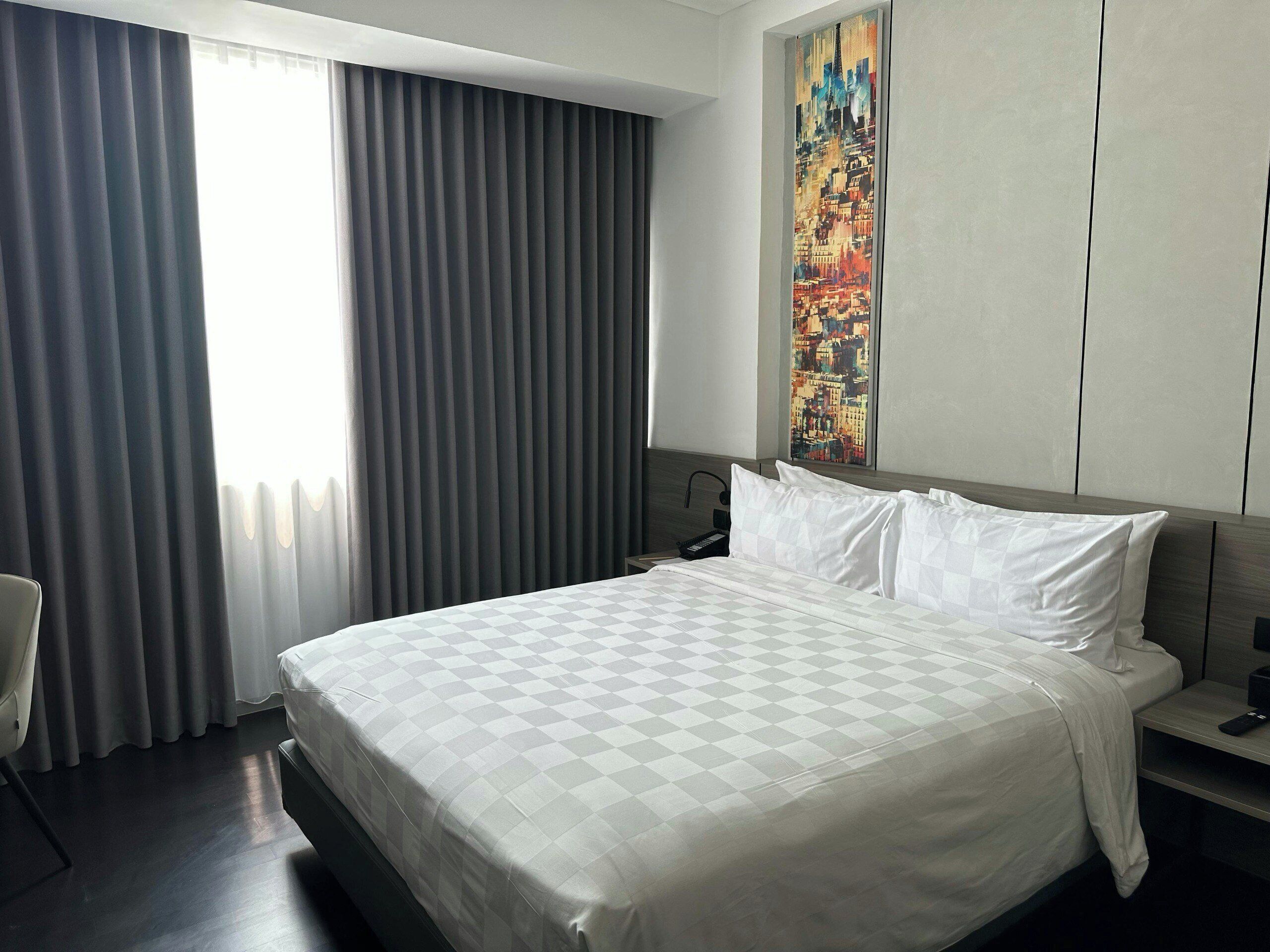 Una habitación de hotel con una cama grande, un cuadro en la pared y una ventana con cortinas.