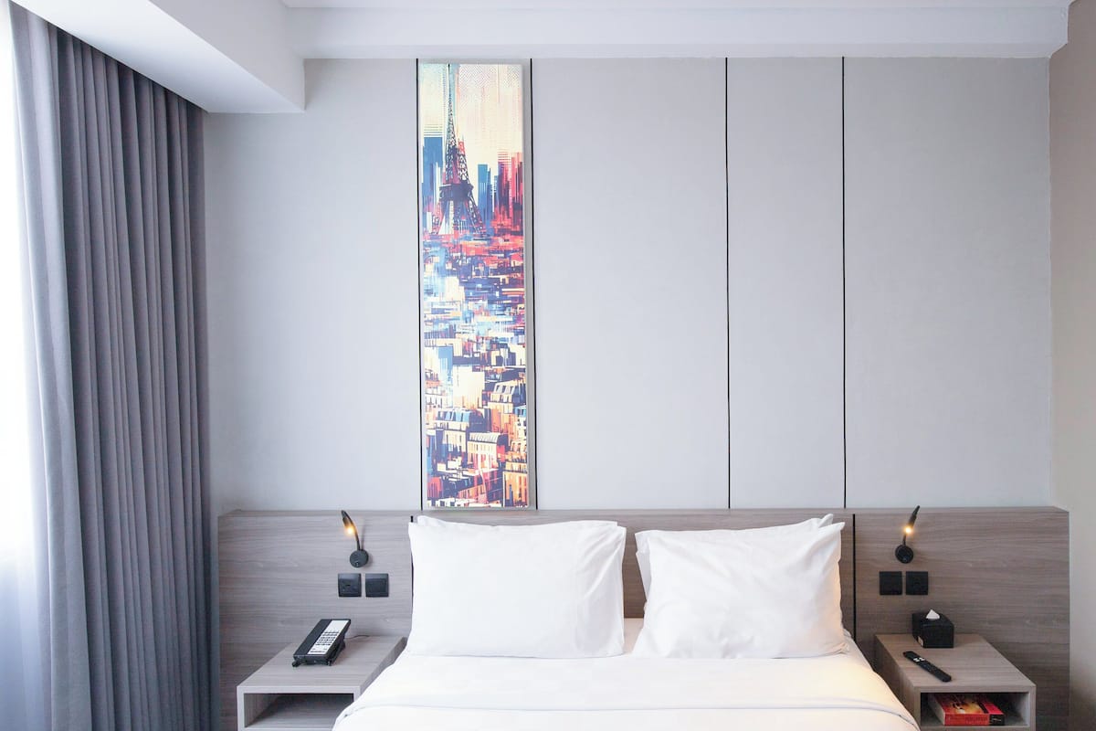 Une chambre d’hôtel avec un grand lit, deux tables de chevet, une œuvre d’art représentant un paysage urbain accrochée au mur et un rideau à la fenêtre.