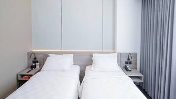 Quarto de hotel com duas camas de solteiro, uma mesa de cabeceira com abajur, um telefone e uma pequena estante com livros.